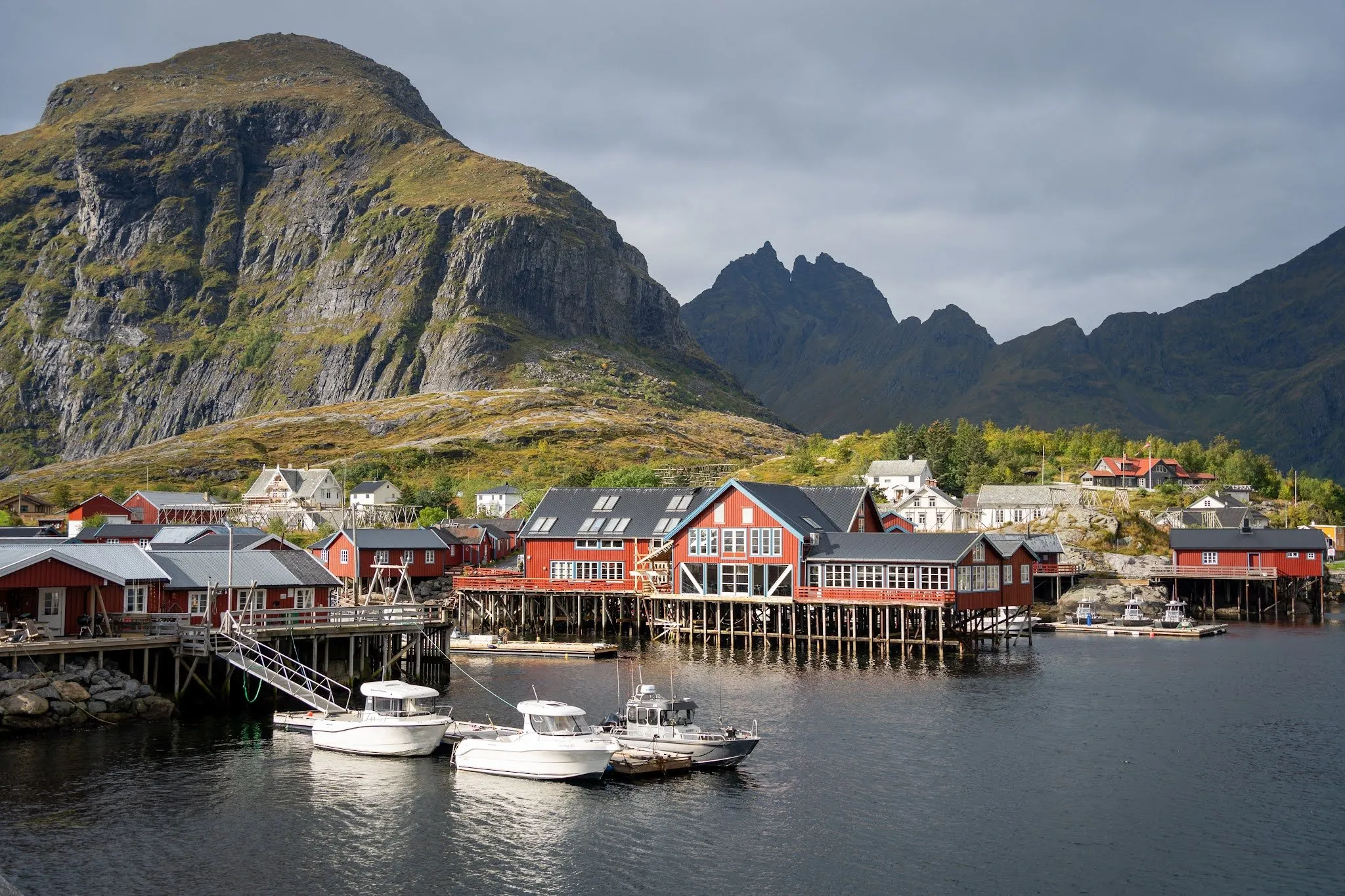 Red fishing houses on stilts and white boats in the harbour of Å, a steep mountain and a jagged mountain ridge behind