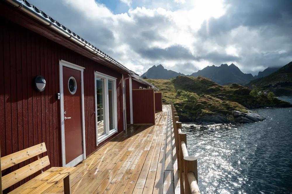 Holzterrasse vor einem der roten Häuschen der Lofoten Cabins Kåkern, rechts das Meer, dahinter Berge unter Wolken