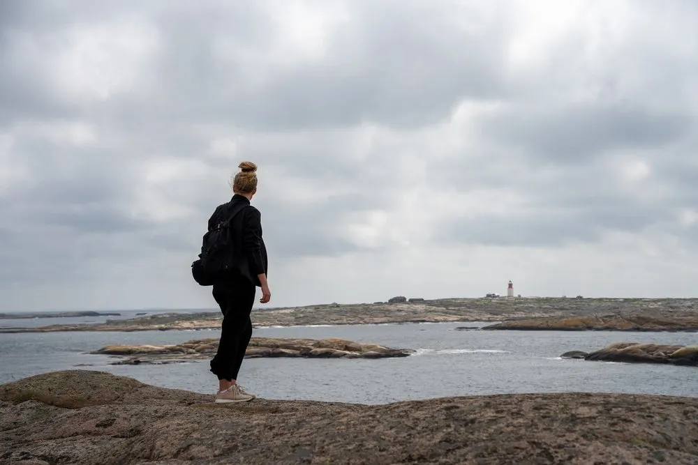 Eine Frau mit Rucksack steht auf dem Fels und blickt aufs Meer, am Horizont ein kleiner Leuchtturm
