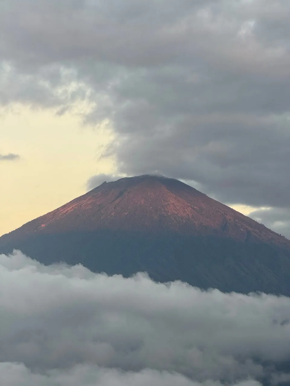 The reddish lit cone of Mount Agung above a band of cloud, grey clouds hanging around the summit