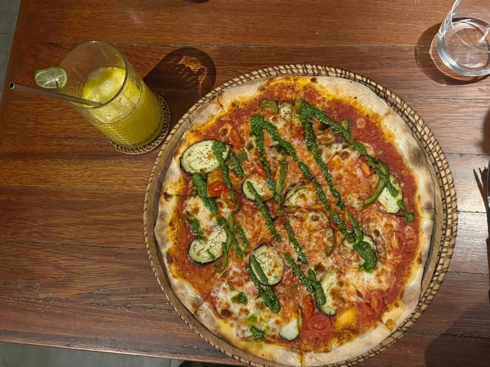 Pizza with tomato sauce, cheese, vegetable slices and green pesto on a woven tray, a yellow drink next to it