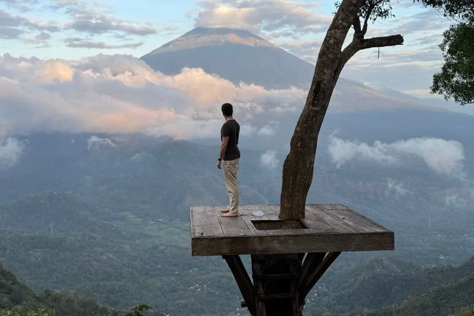Nicolas stands barefoot on a wooden platform around a tree and looks across a valley with pink-lit clouds at the volcano Mount Agung
