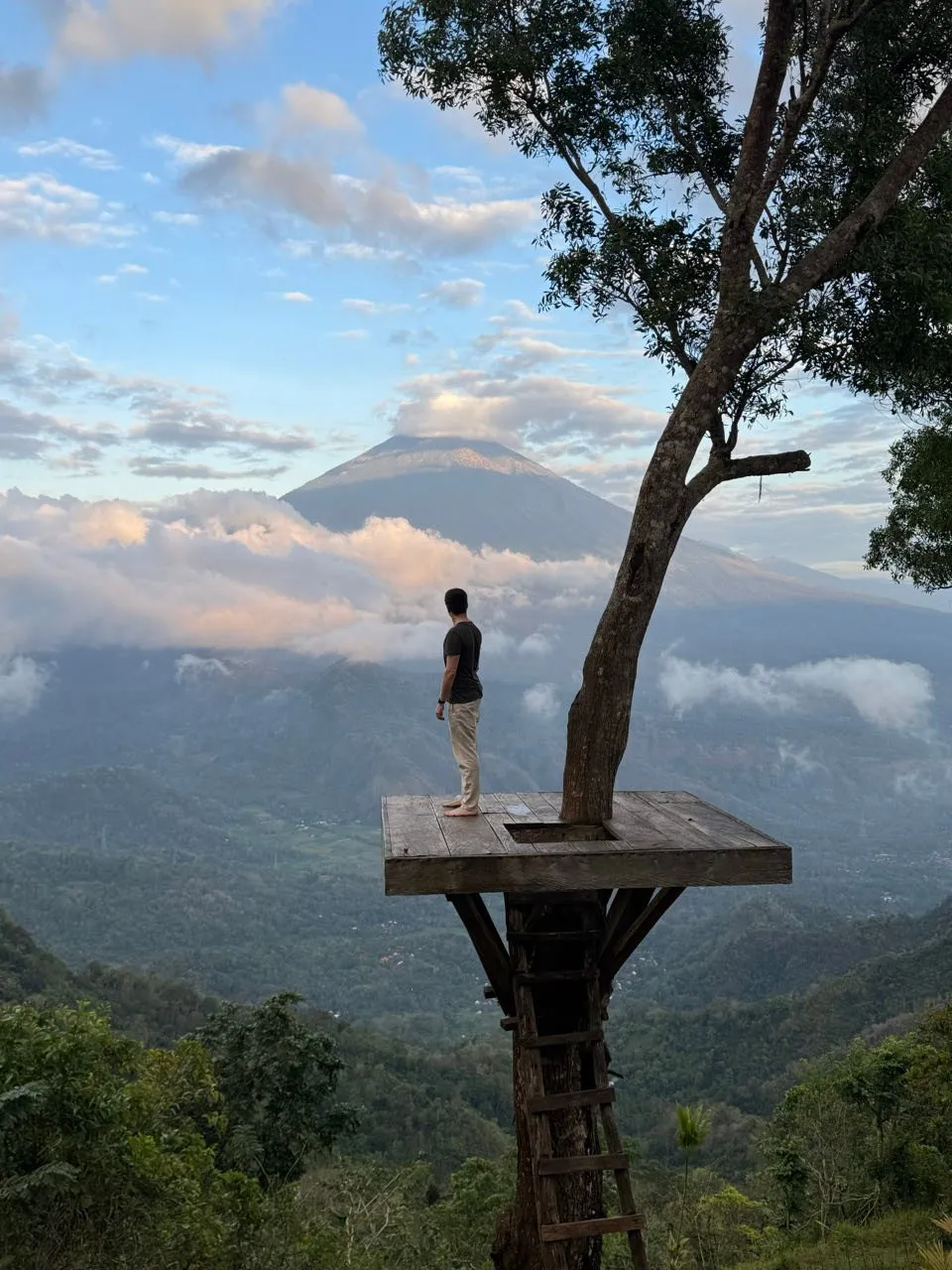 Nicolas stands barefoot on a wooden platform around a tree, the volcano rising behind the valley with its clouds