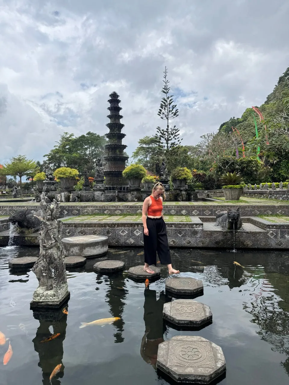 Annina balances barefoot across stepping stones in the koi pond of Tirta Gangga, a multi-tiered tower behind her