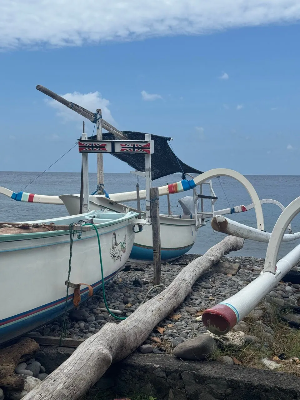 Zwei Auslegerboote mit bunten Streifen auf dunklen Kieselsteinen am Strand, an einem davon zwei Union-Jack-Flaggen, dahinter das blaue Meer