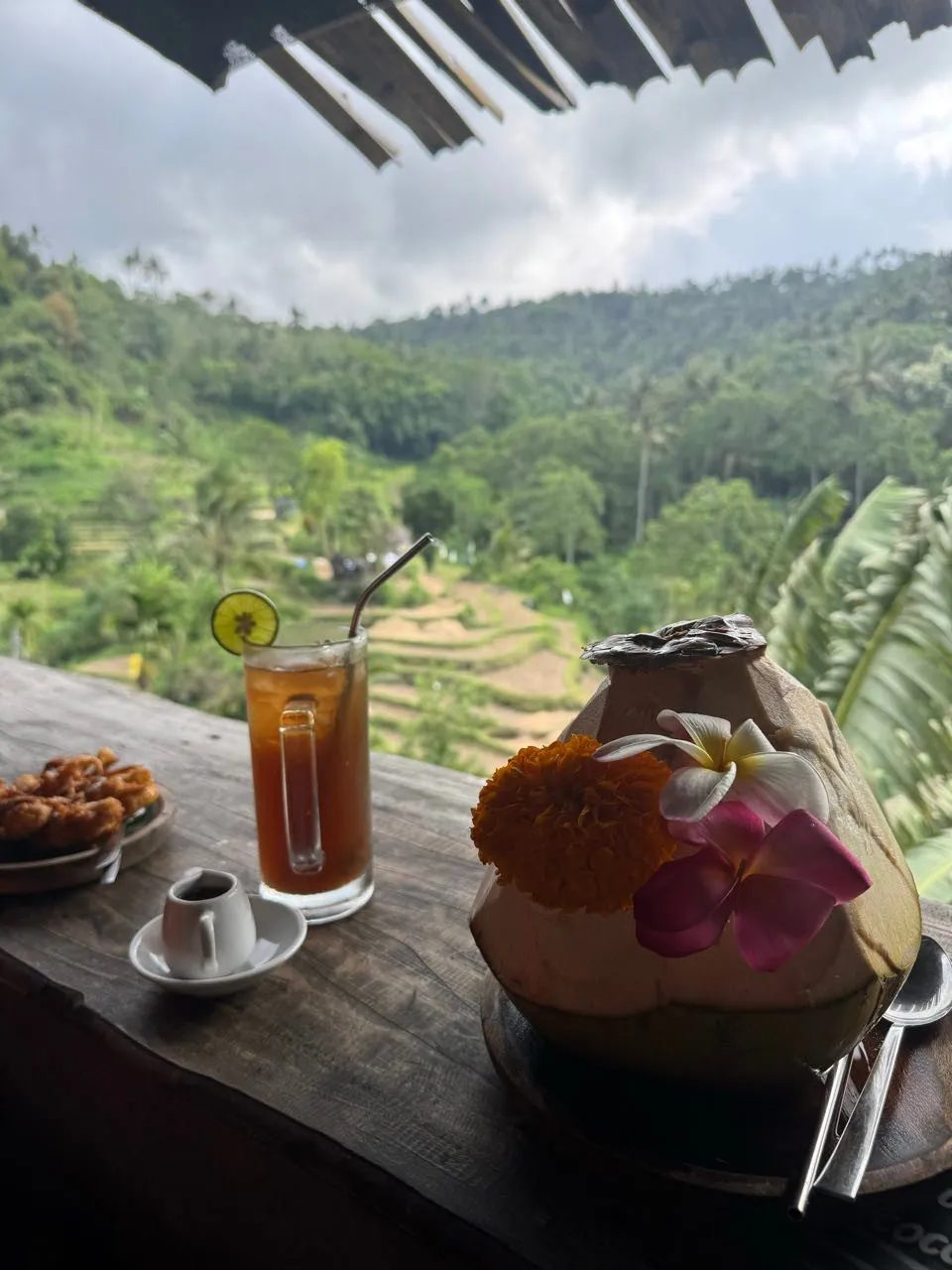 On the wooden table stand fried banana, a glass with a slice of lime and a coconut decorated with flowers, the terraces behind