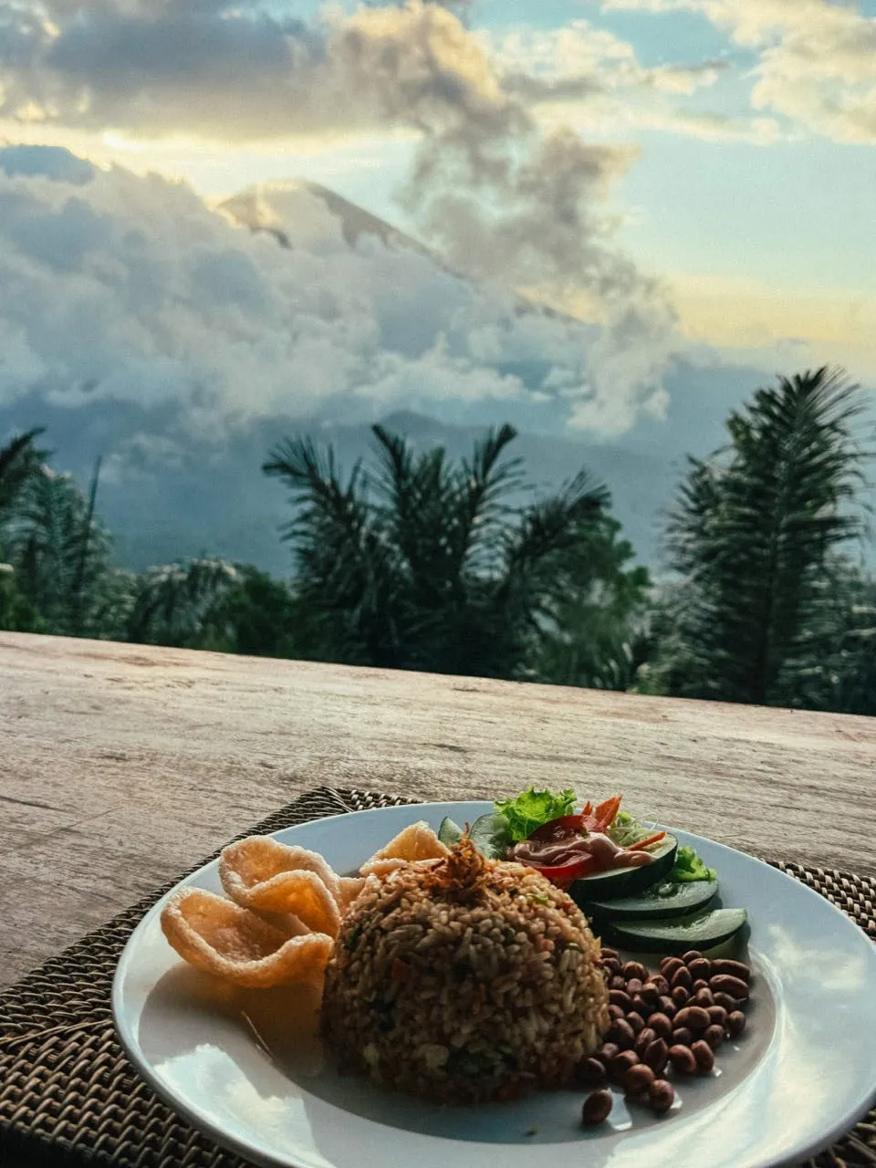 A plate of fried rice, krupuk and cucumber slices, with clouds and a mountain peak in the evening light behind