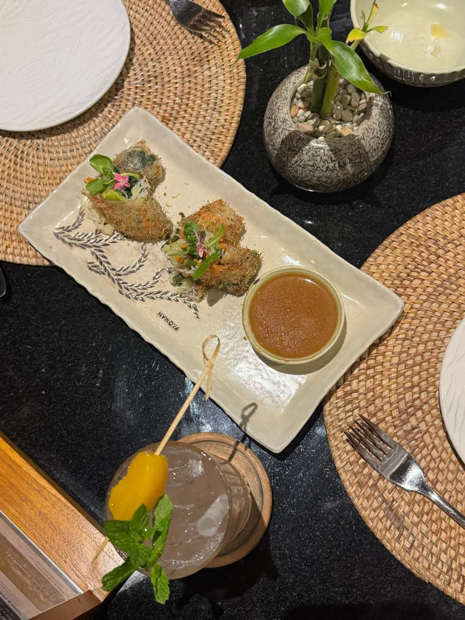 Zwei knusprige Röllchen mit Kräutern und Blüten, daneben Dipsauce und ein Glas mit Minze und Fruchtspiess