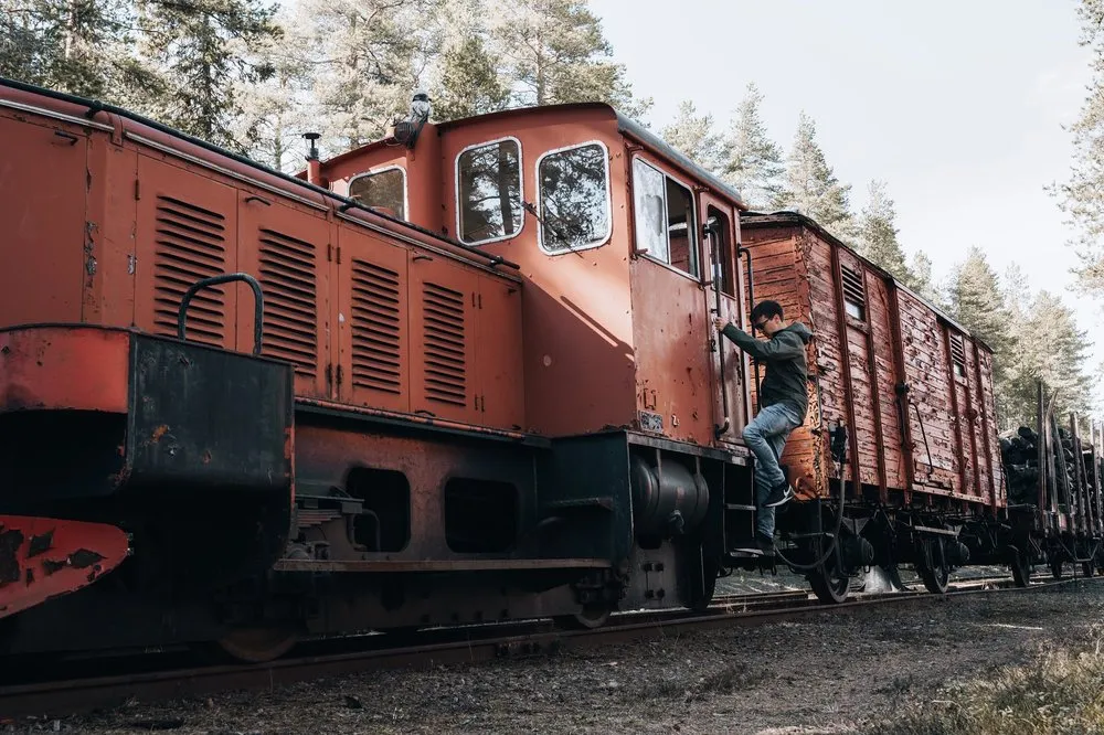 A man climbs up the side of the red locomotive, freight cars and pines behind it