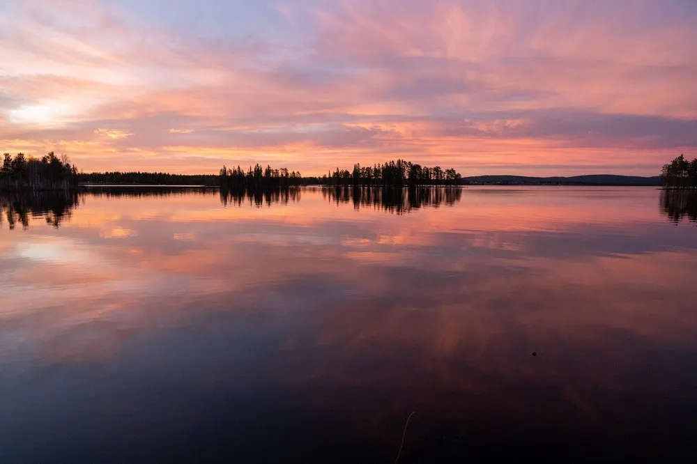 Pink and orange clouds are reflected in the mirror-smooth lake, tree silhouettes stand on the shore