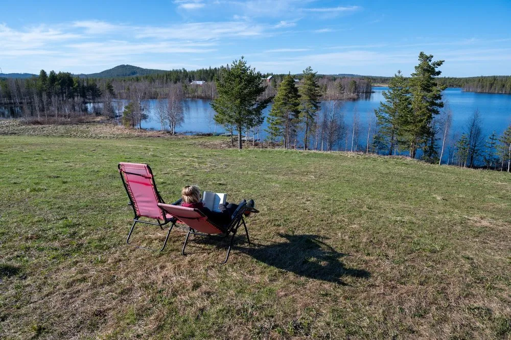 A person reads in the red deckchair, behind the meadow lies the lake with wooded shores