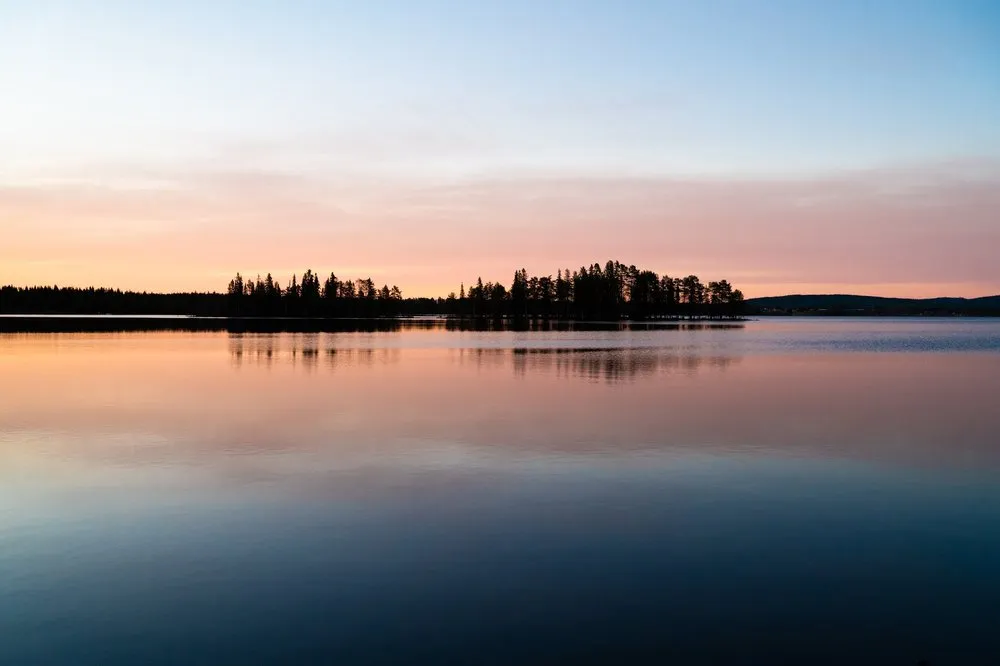 Pale evening sky in pink and blue, a wooded headland reflected in the calm lake