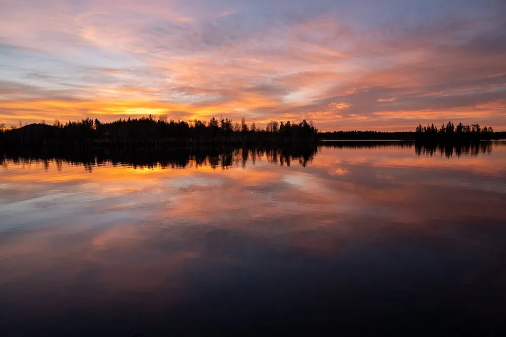 Orange and violet clouds above the lake, the row of trees on the shore reflected in the dark water