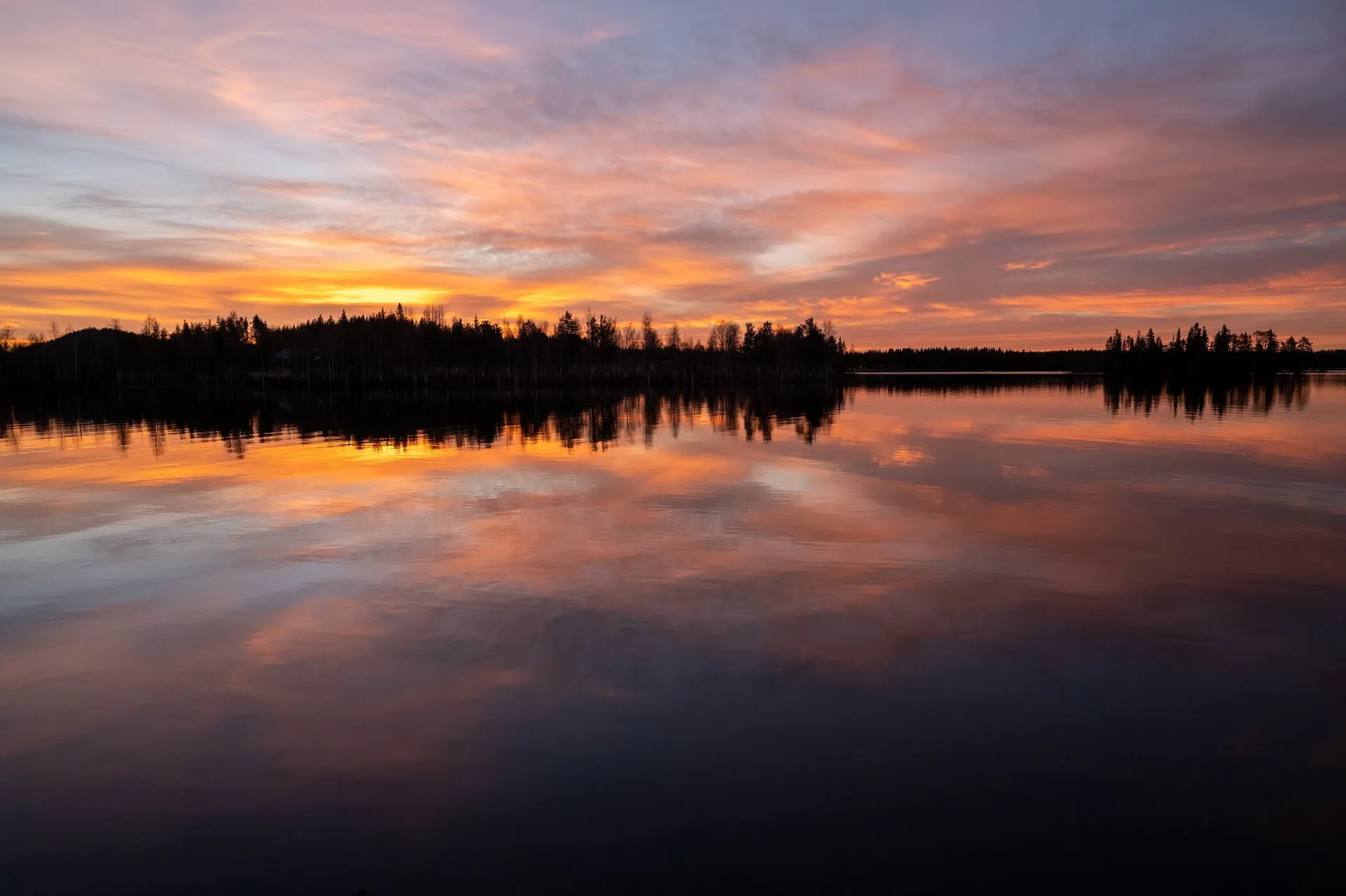 Orange und violette Wolken über dem See, die Baumreihe am Ufer spiegelt sich im dunklen Wasser