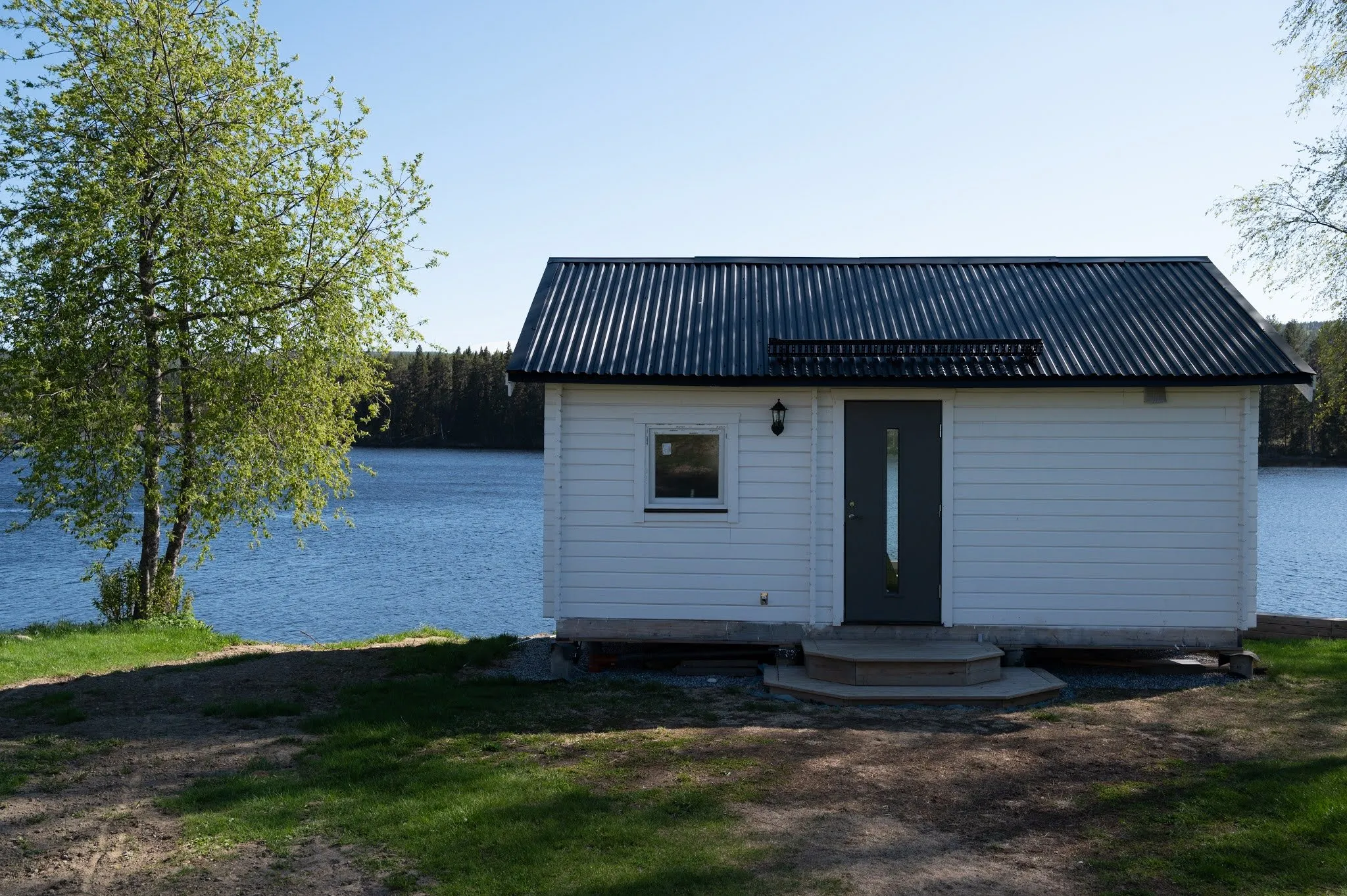 A small white cottage with a dark roof stands on a meadow right by the blue lake
