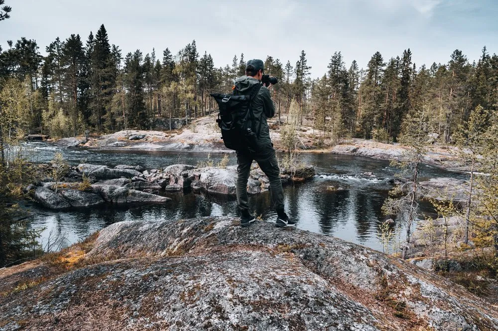 Ein Mann mit Kamera und Rucksack steht auf einem Granitfelsen über einem Fluss mit Stromschnellen, dahinter Kiefernwald