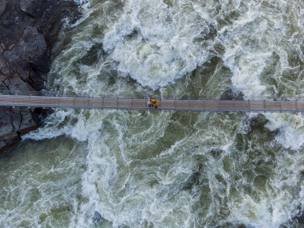 Vertical aerial shot of the suspension bridge over the swirling water of the Handölsforsen, two small people in the middle