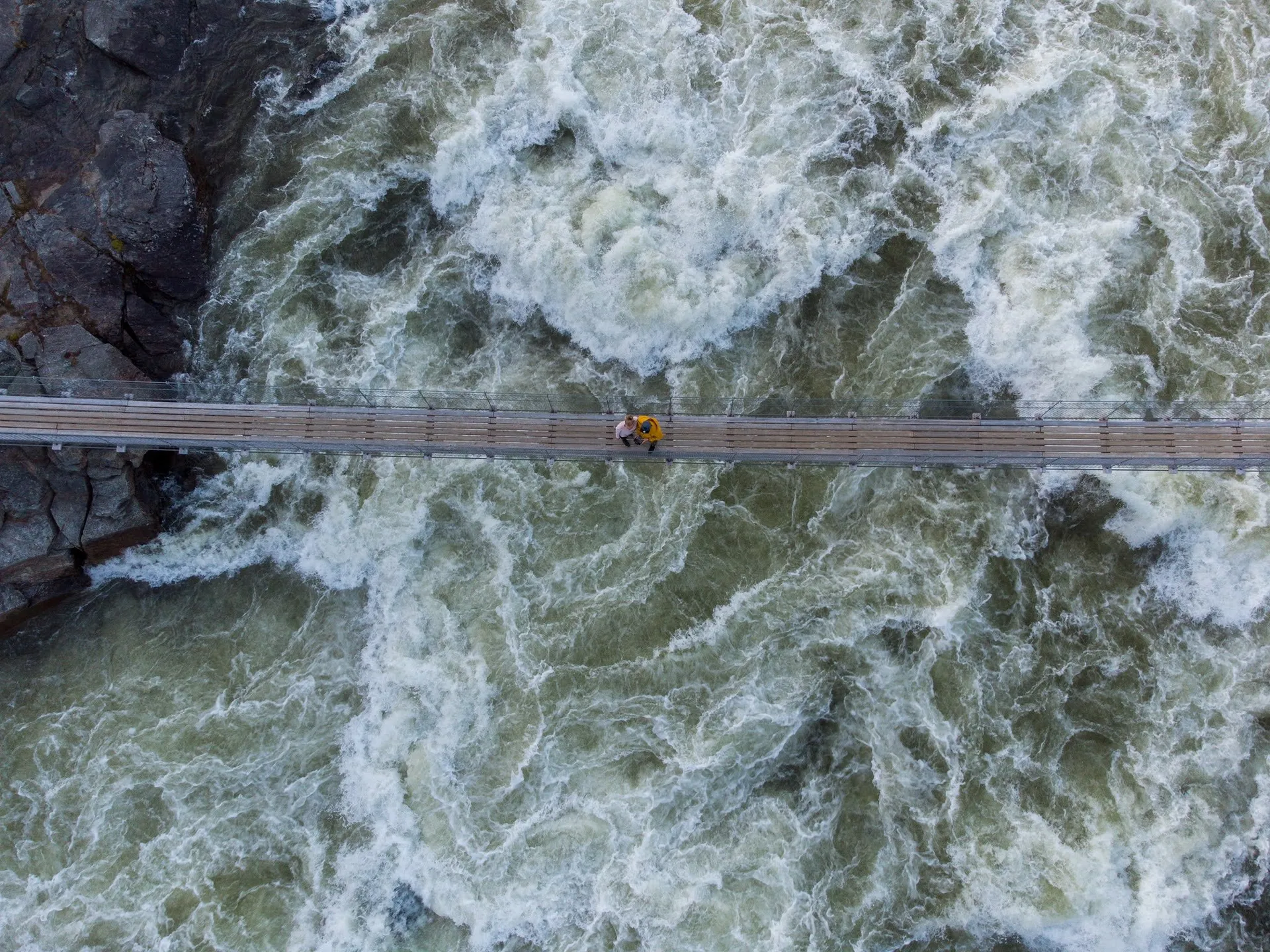 Senkrechte Luftaufnahme der Hängebrücke über dem wirbelnden Wasser des Handölsforsen, in der Mitte zwei kleine Personen