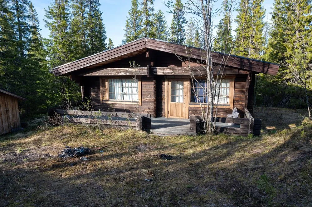 The dark brown log cabin of the Airbnb in the forest with a veranda, a small wooden shed on the left