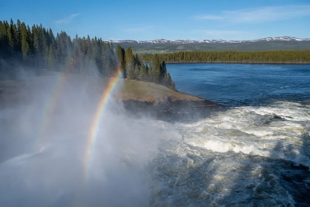 Double rainbow in the spray of the Tännforsen, forest, a lake and mountains with patches of snow behind
