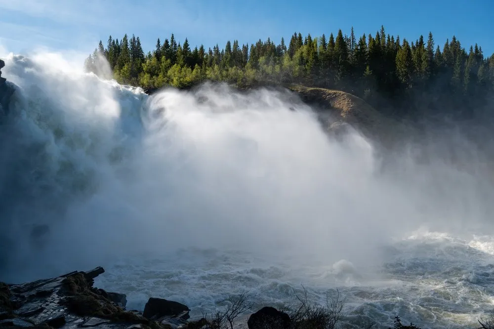 A huge cloud of spray over the Tännforsen, a strip of conifer forest under a blue sky above