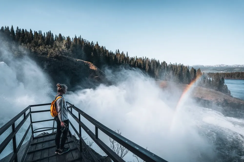 Eine Person steht auf einer Holzplattform mit Geländer in der Gischt des Tännforsen, rechts leuchtet ein Regenbogen