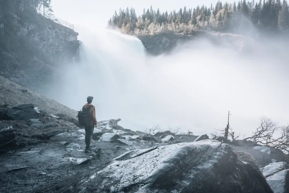 A man with a backpack stands on wet rocks in front of the wall of spray of the Tännforsen