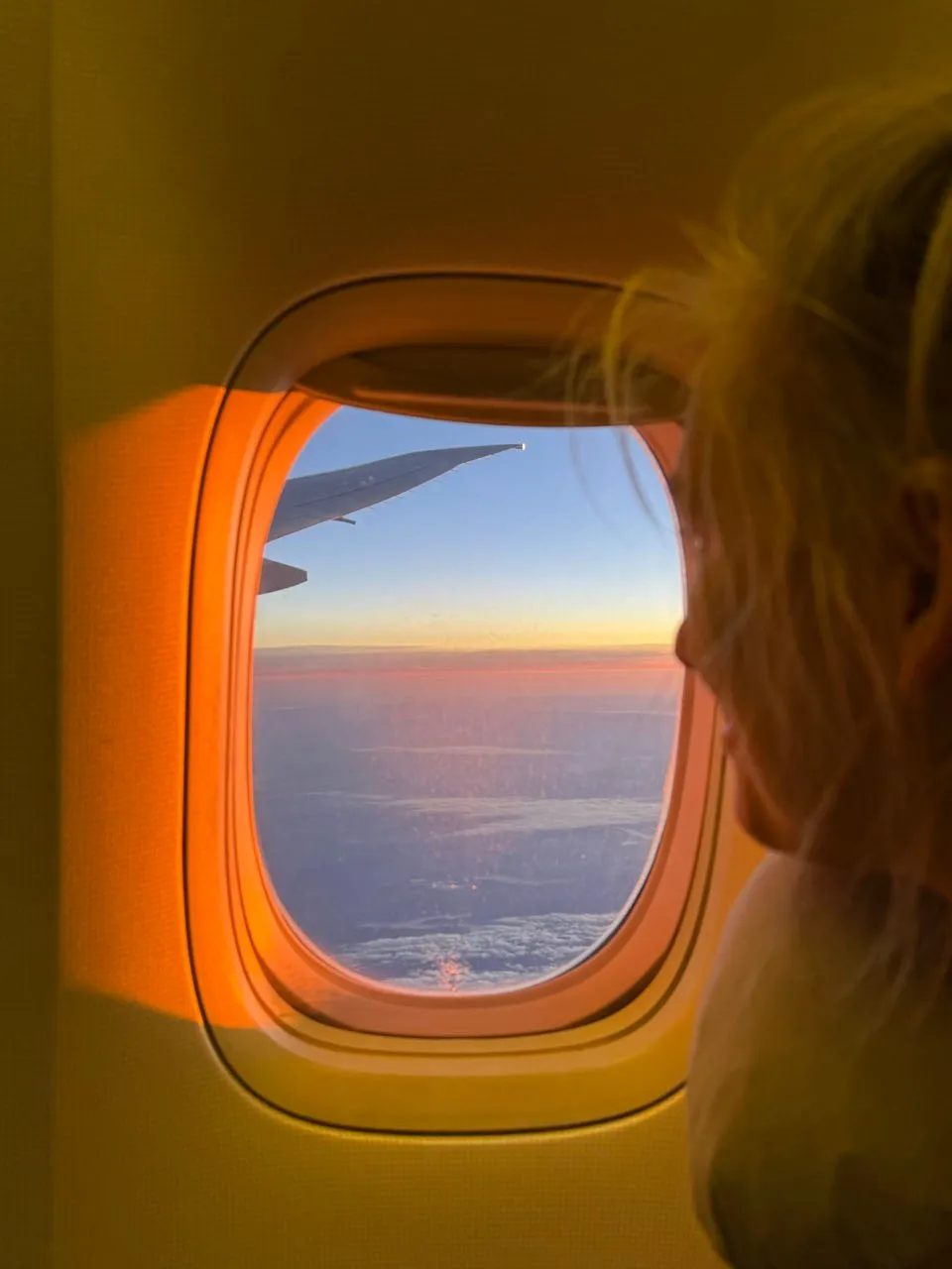 Blick aus dem Flugzeugfenster auf ein Wolkenmeer mit orangem Horizont, im Anschnitt eine Person mit blonden Haaren