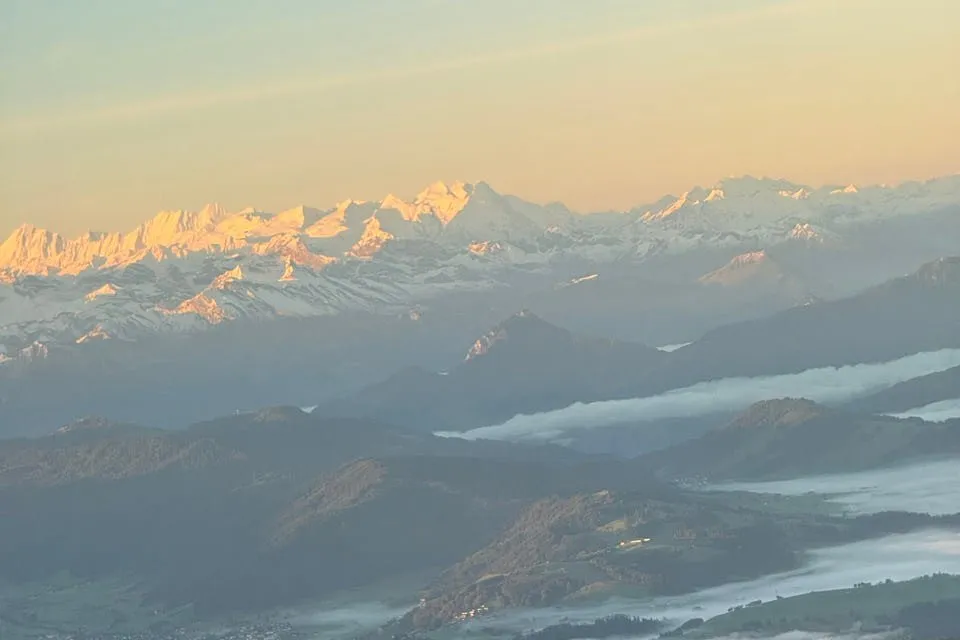 Orange-lit snowy peaks of the Alps above dark hills and bands of fog in the morning light