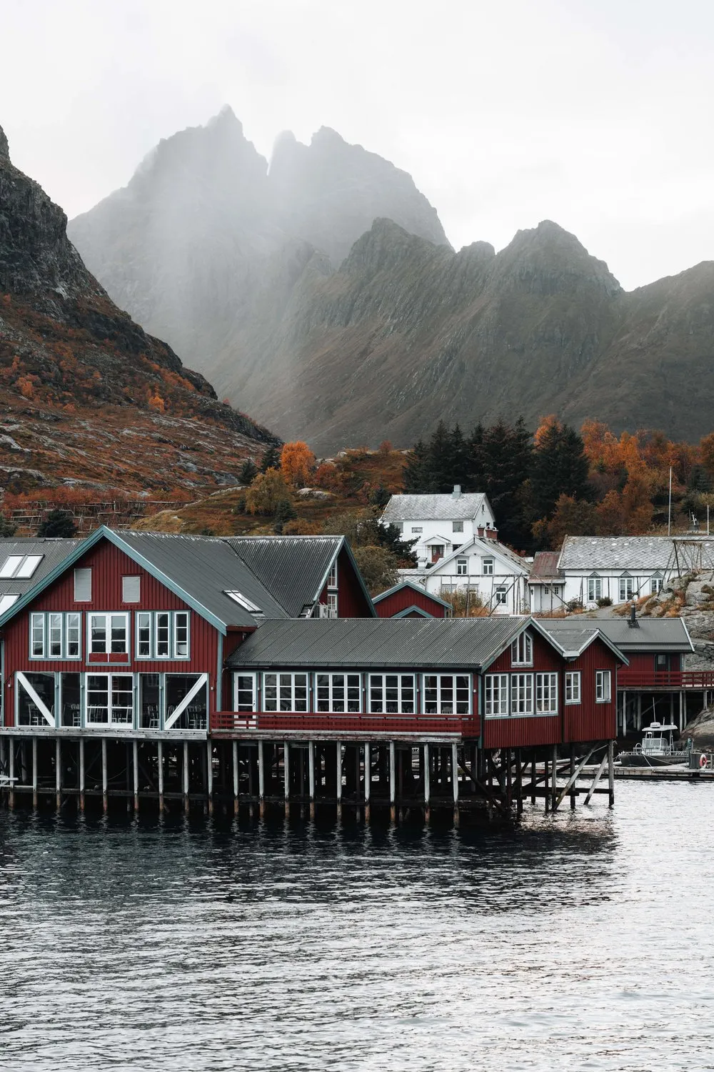 Rote Rorbuer auf Stelzen am Wasser in Å, dahinter herbstliche Bäume und Berge im Nebel