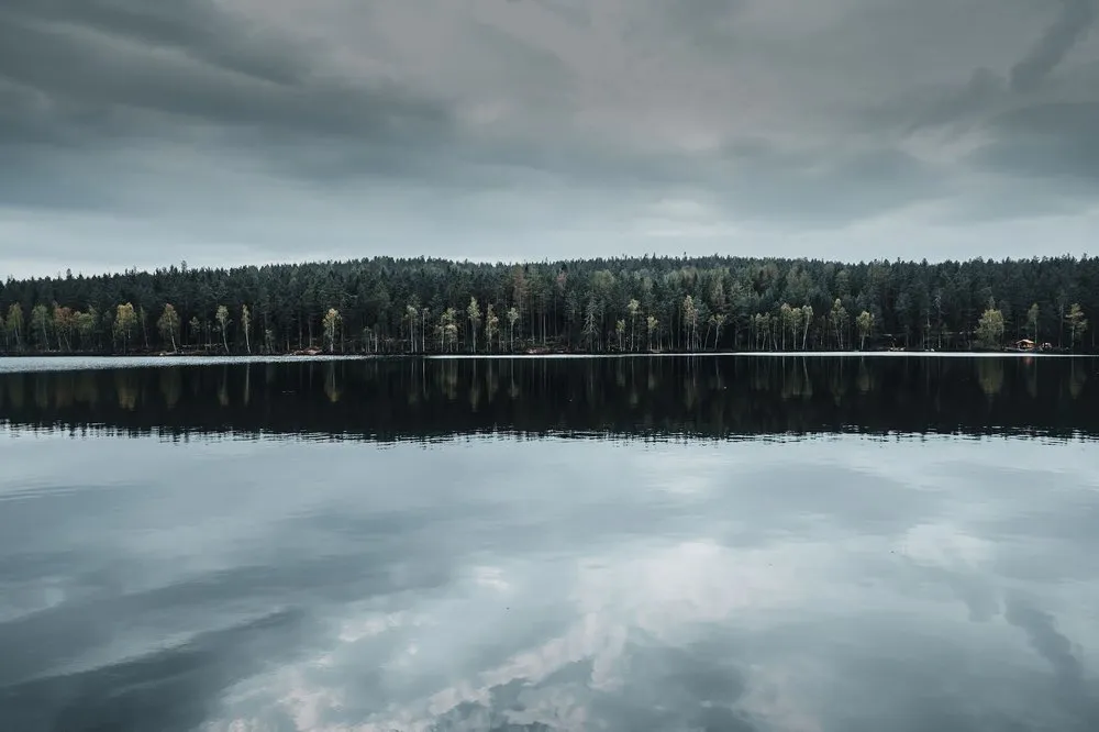 Dunkle Wolken über einem Waldsee, das bewaldete Ufer spiegelt sich im ruhigen Wasser