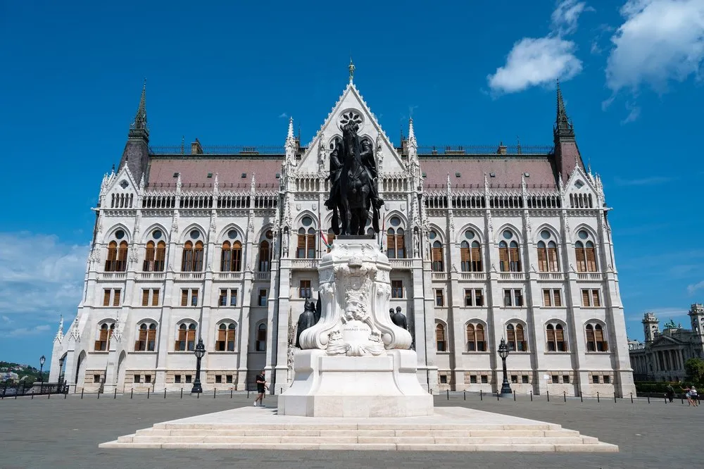 Front view of the Parliament building with an equestrian statue on a light plinth, in front of it a wide square under a blue sky