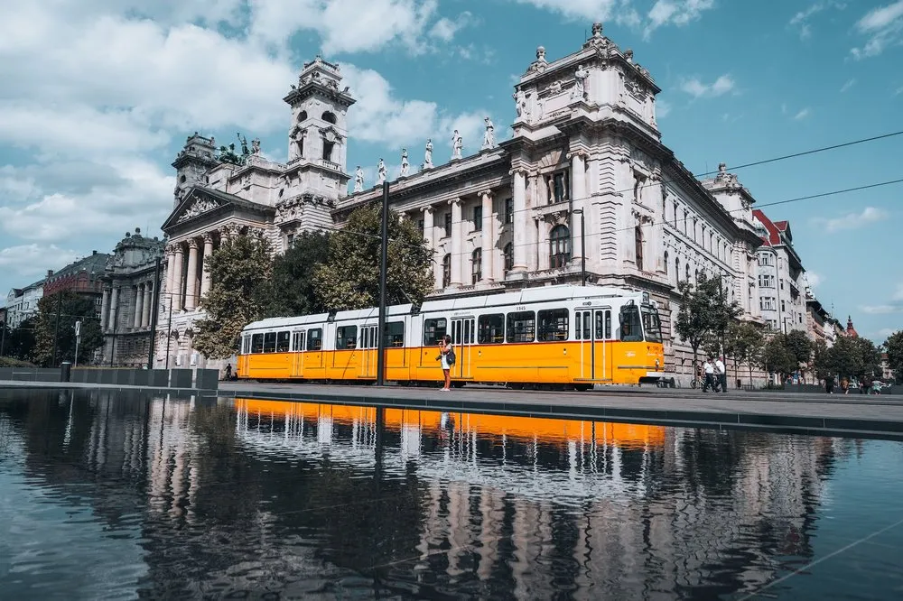 A yellow tram passes a light-coloured building with a tower, which is reflected in the water in front of it