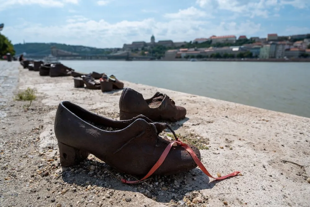 Ein dunkler Schuh mit rotem Band im Vordergrund, dahinter weitere Schuhe am Rand der Ufermauer und die Donau