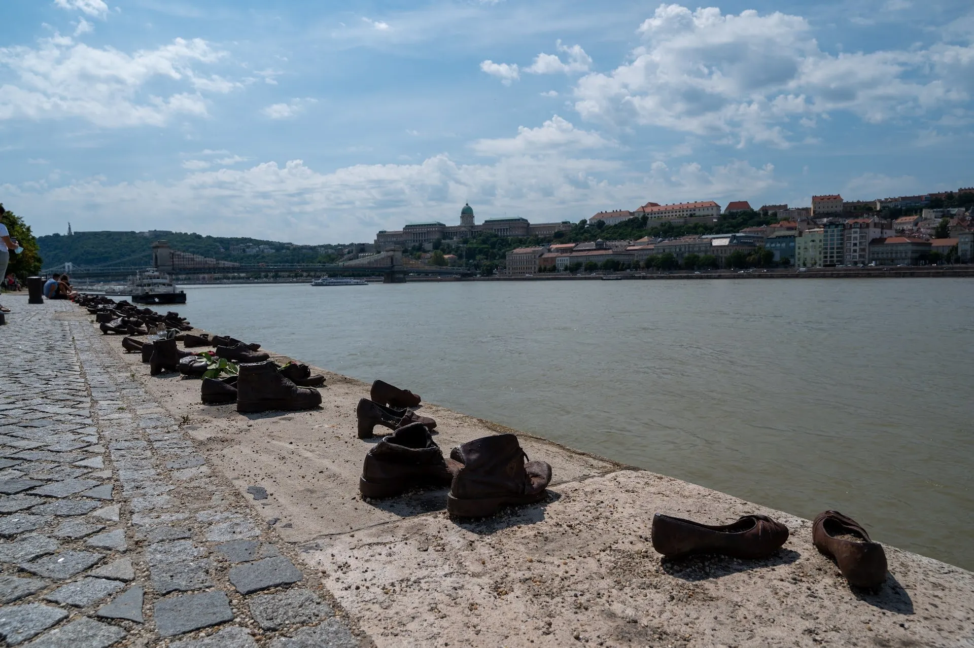 A row of shoes made of dark metal on the stone edge of the Danube bank, a bridge and a domed building in the background