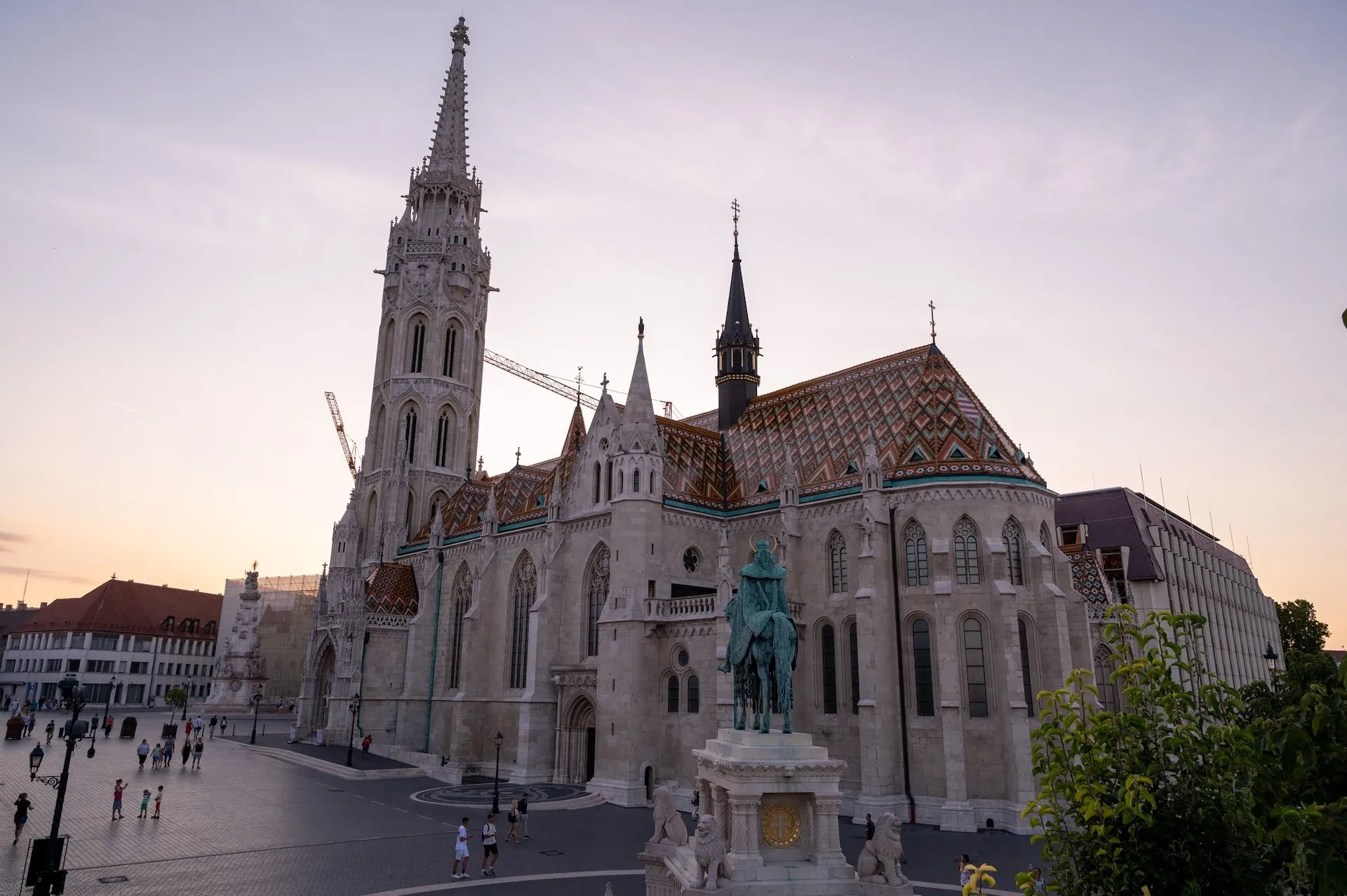 The church with a tall tower and a colourful tiled roof in the evening light, in front of it an equestrian statue on the square