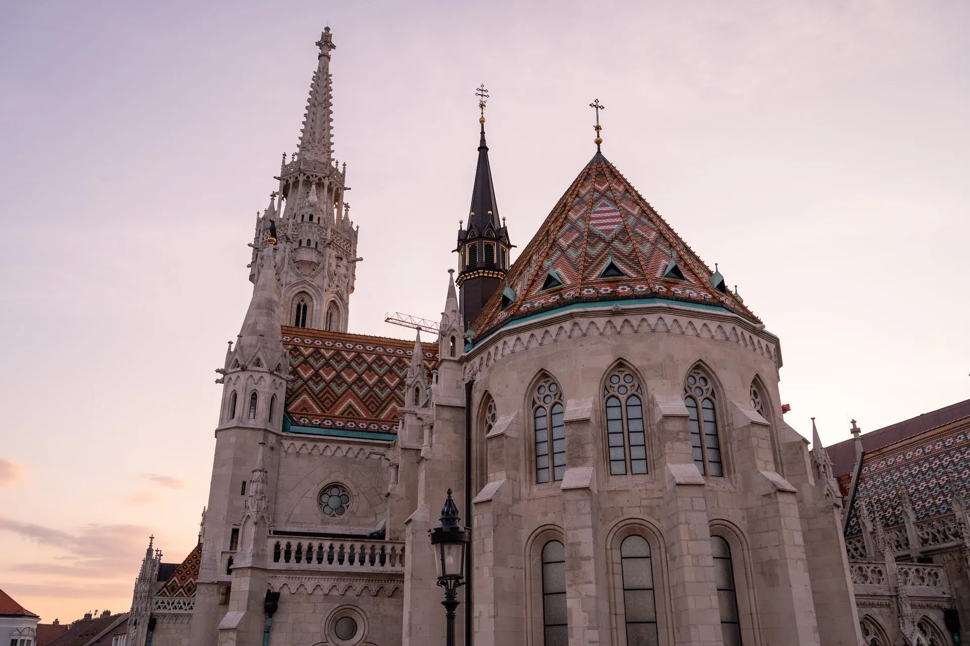 The patterned tiled roof and the tower of the church from below against a pink sky, a street lamp in front