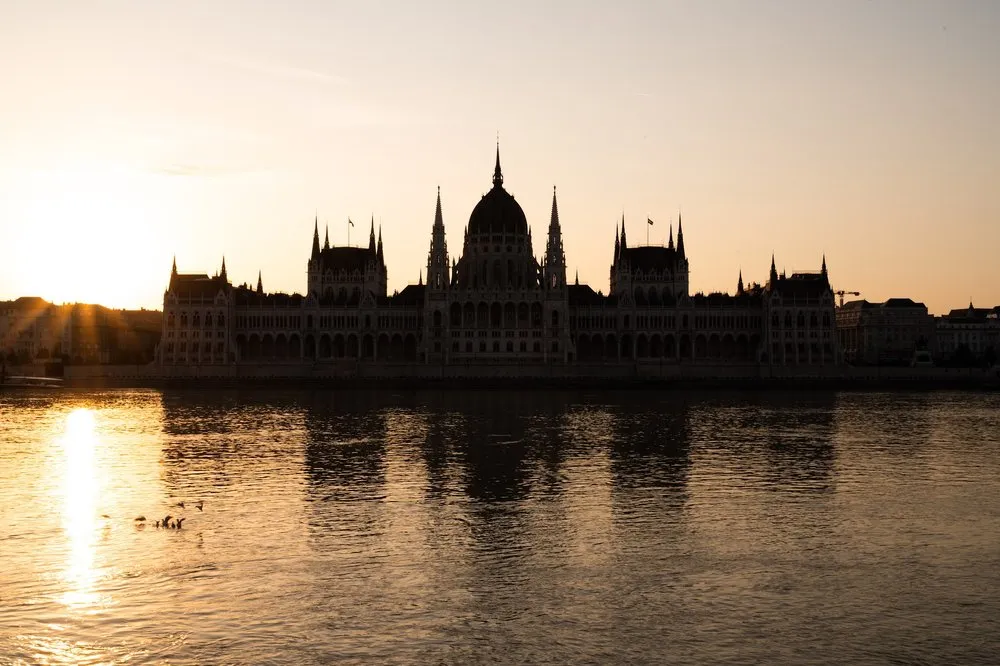Das Parlament als dunkle Silhouette vor dem hellen Morgenhimmel, links spiegelt sich die Sonne im ruhigen Wasser der Donau