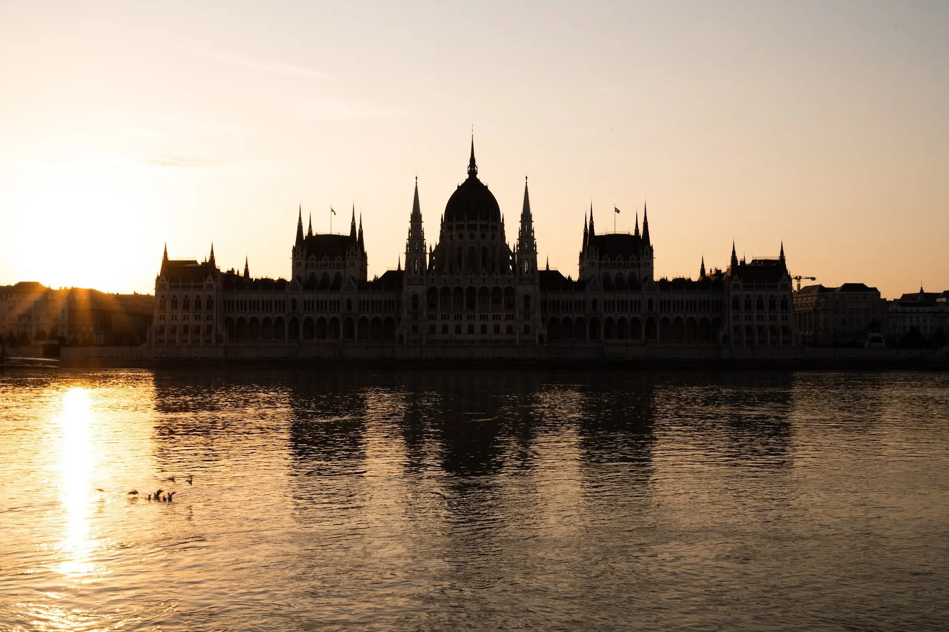 The Parliament as a dark silhouette against the bright morning sky, on the left the sun is reflected in the calm water of the Danube