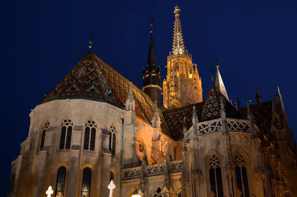 Eine angestrahlte Kirche mit buntem Ziegeldach und spitzem Turm vor dem dunkelblauen Nachthimmel