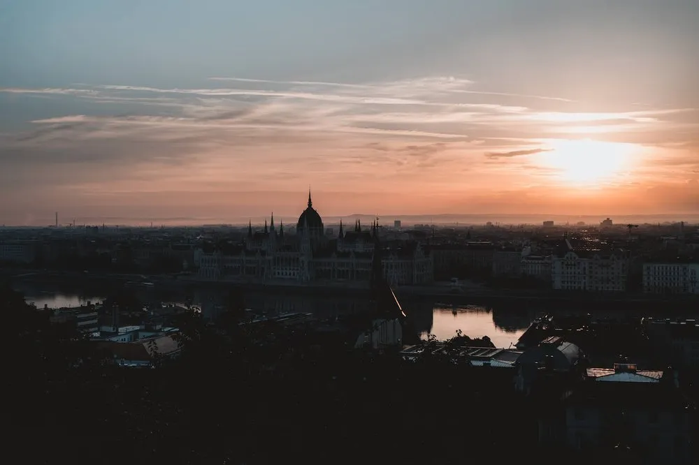 Das Parlament als Silhouette im Morgenrot, rechts die Sonne über dem Dunst und davor der Fluss