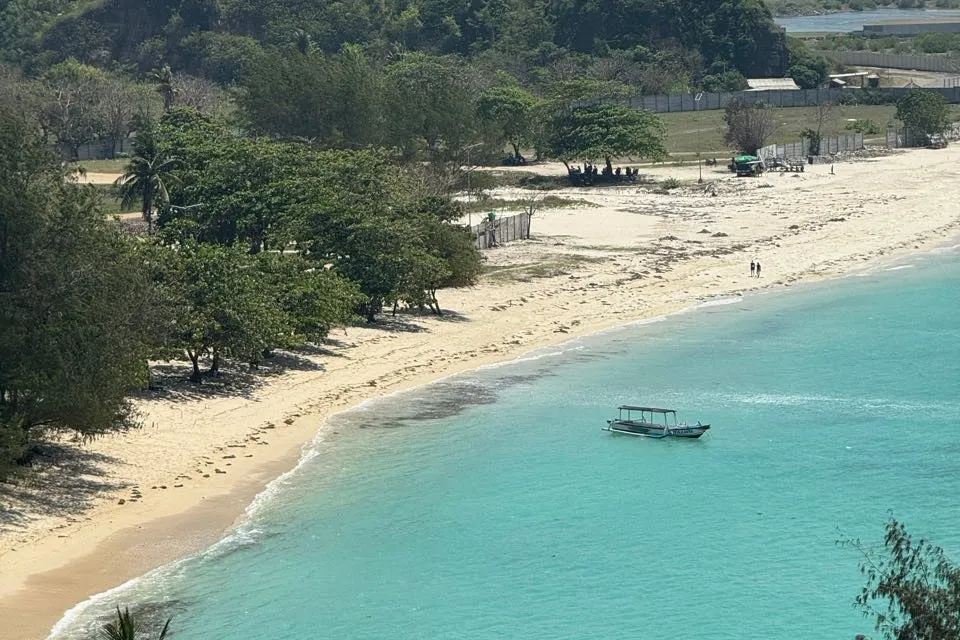 A small boat lies in turquoise water in front of a light sandy beach with trees