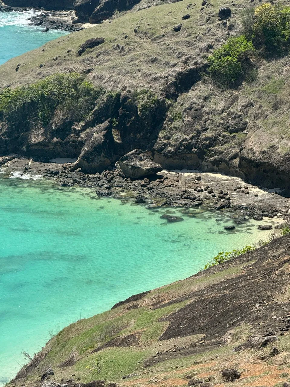 Small rocky bay with turquoise water, dark rocks, a narrow strip of sand and a grassy slope