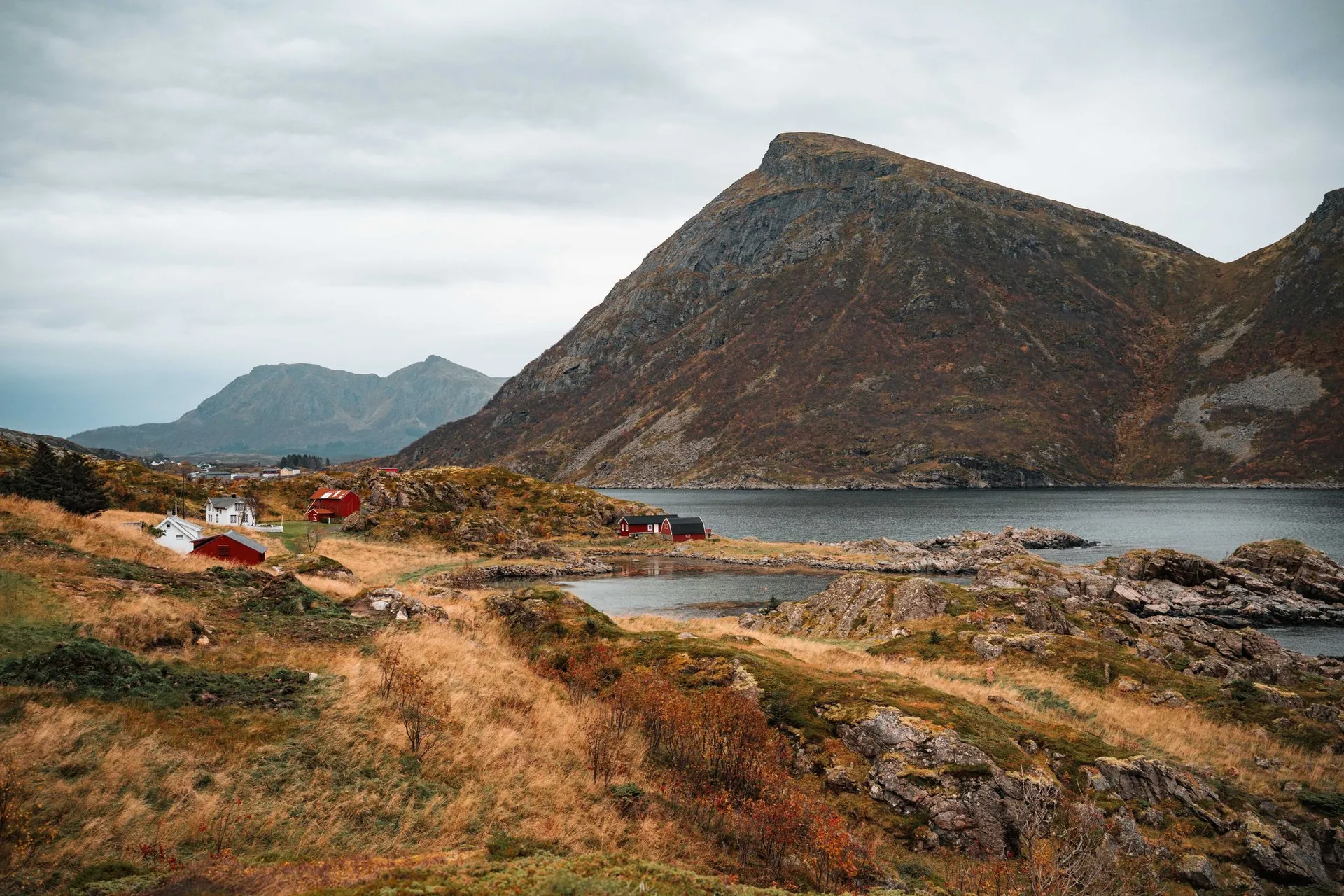 Rote und weisse Häuser an einer Bucht unter einem spitzen Berg, vorne Herbstgras und Felsen am Coastal Trail