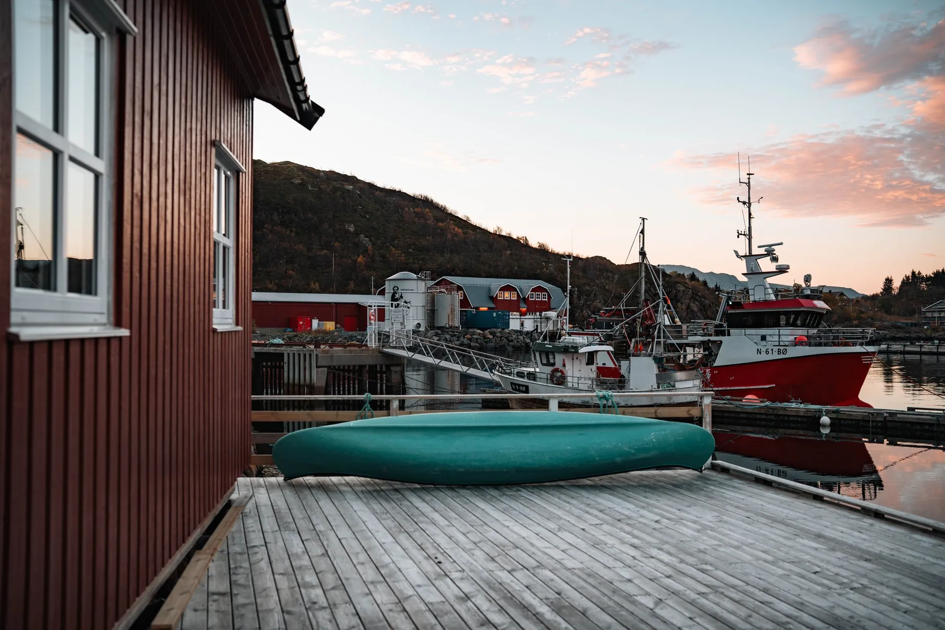 Ein grünes Kajak auf einem Holzsteg neben einem roten Holzhaus, dahinter Fischerboote im Hafen von Straumsjøen