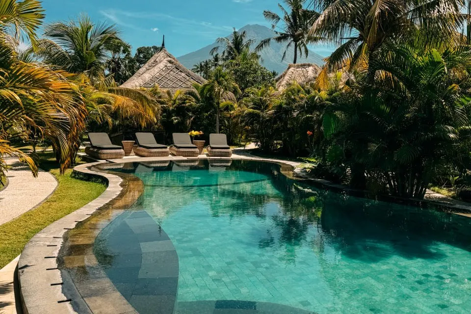 Turquoise pool between palm trees and thatched huts, with a mountain behind
