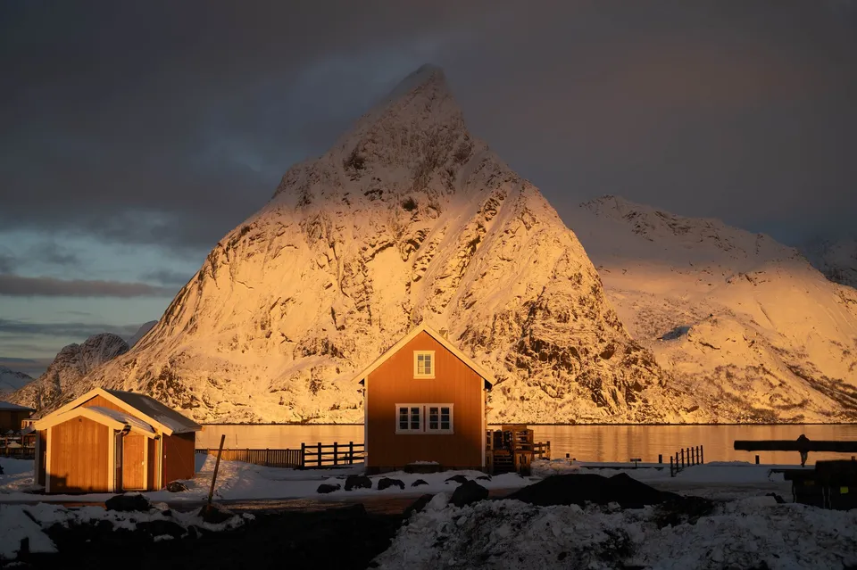 Der verschneite Olstind glüht im ersten Sonnenlicht golden über den Rorbuer von Sakrisøy