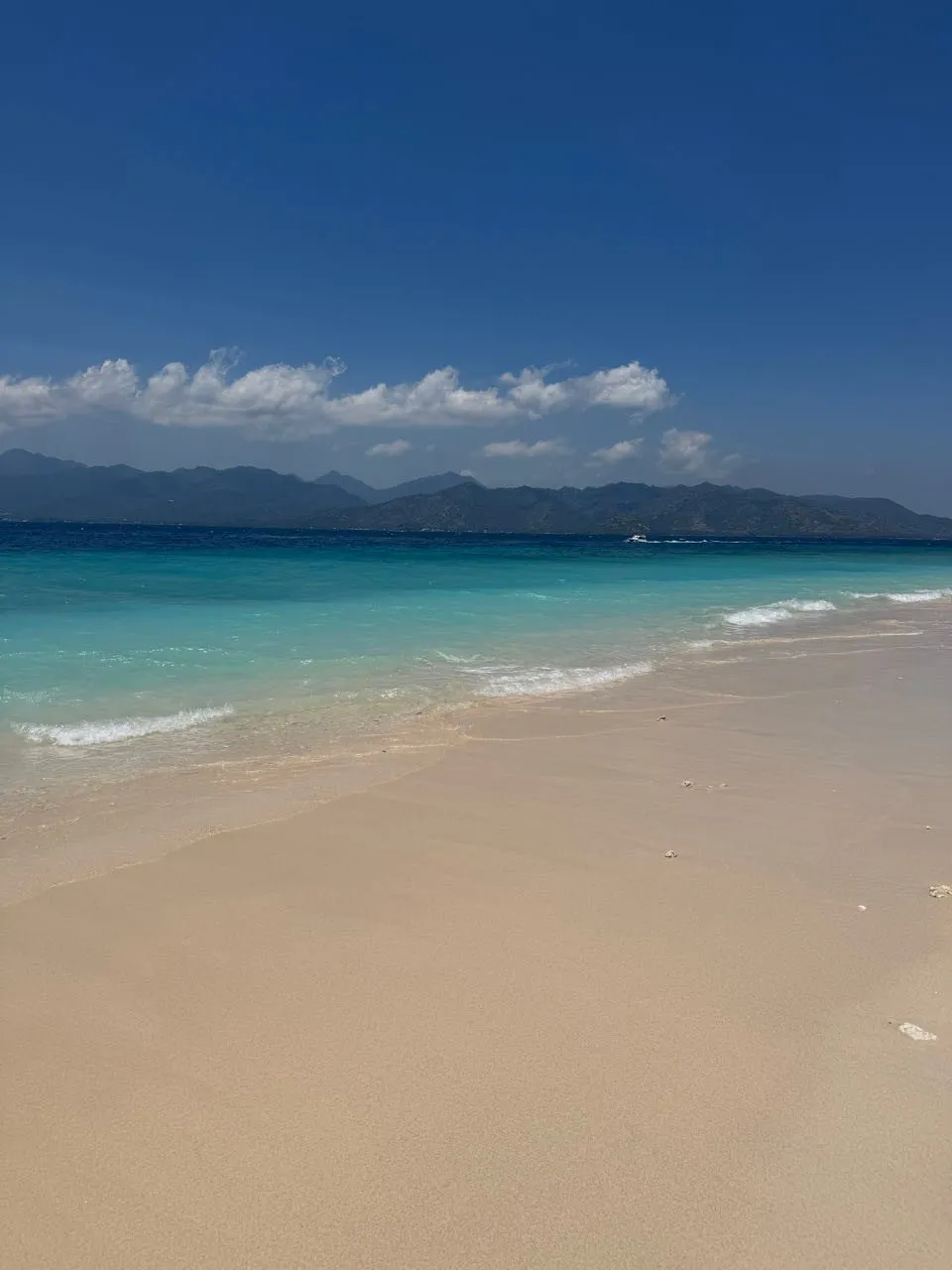 Light sandy beach with gentle waves and turquoise water, dark mountains on the horizon under a blue sky