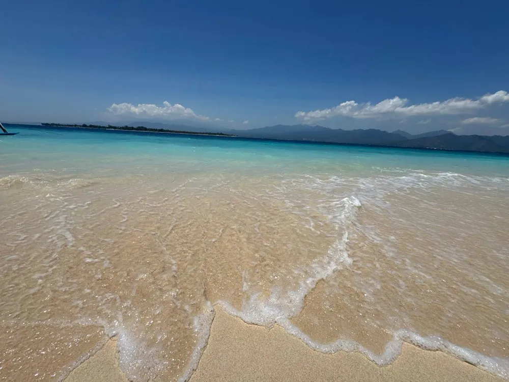 Waves run over light sand into the turquoise water, in the distance an island and mountains