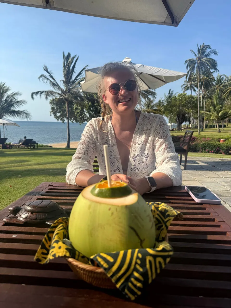 Annina with sunglasses smiles at a wooden table, in front of her a green coconut with a straw, behind her lawn, palm trees and sea
